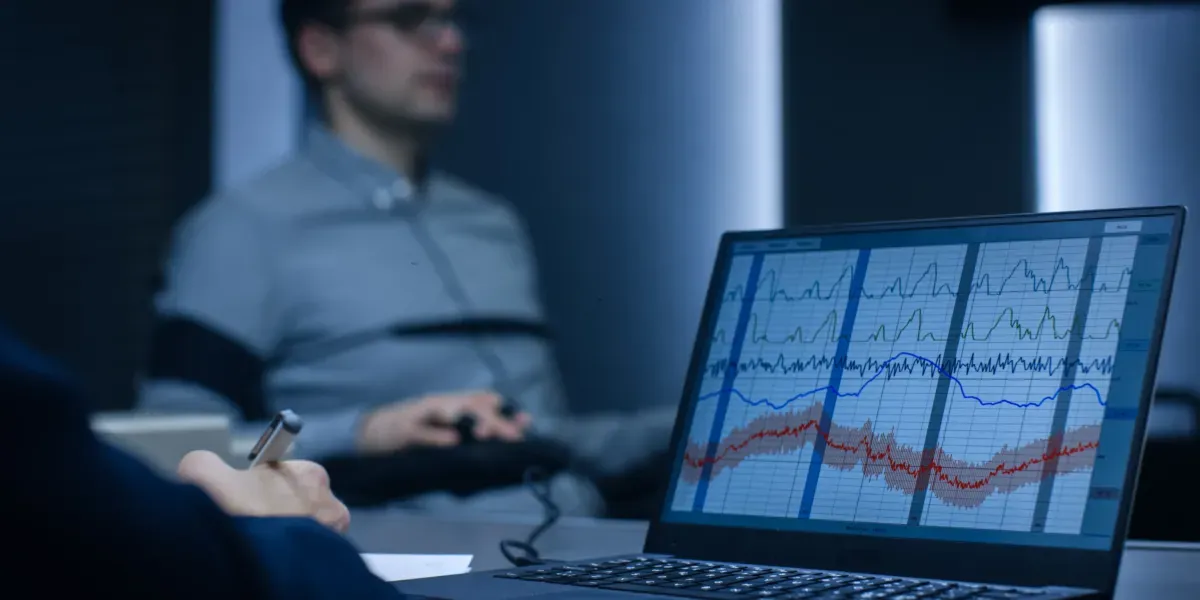A person undergoes a polygraph test, data displayed on a laptop screen. Investigator takes notes.