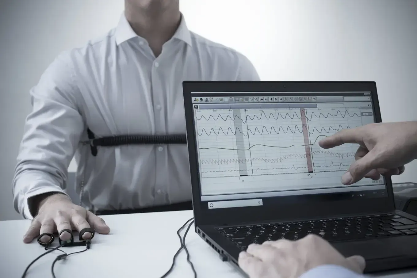 Person undergoing a lie detector test. Wires connect to fingers and chest, with a laptop displaying data and a hand pointing at the screen.