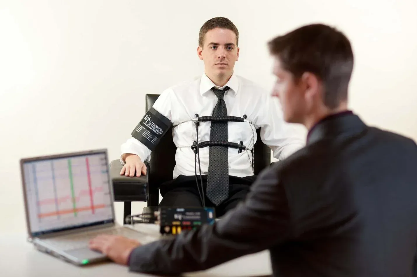 Man connected to a polygraph machine, facing another man with a laptop. Office setting.