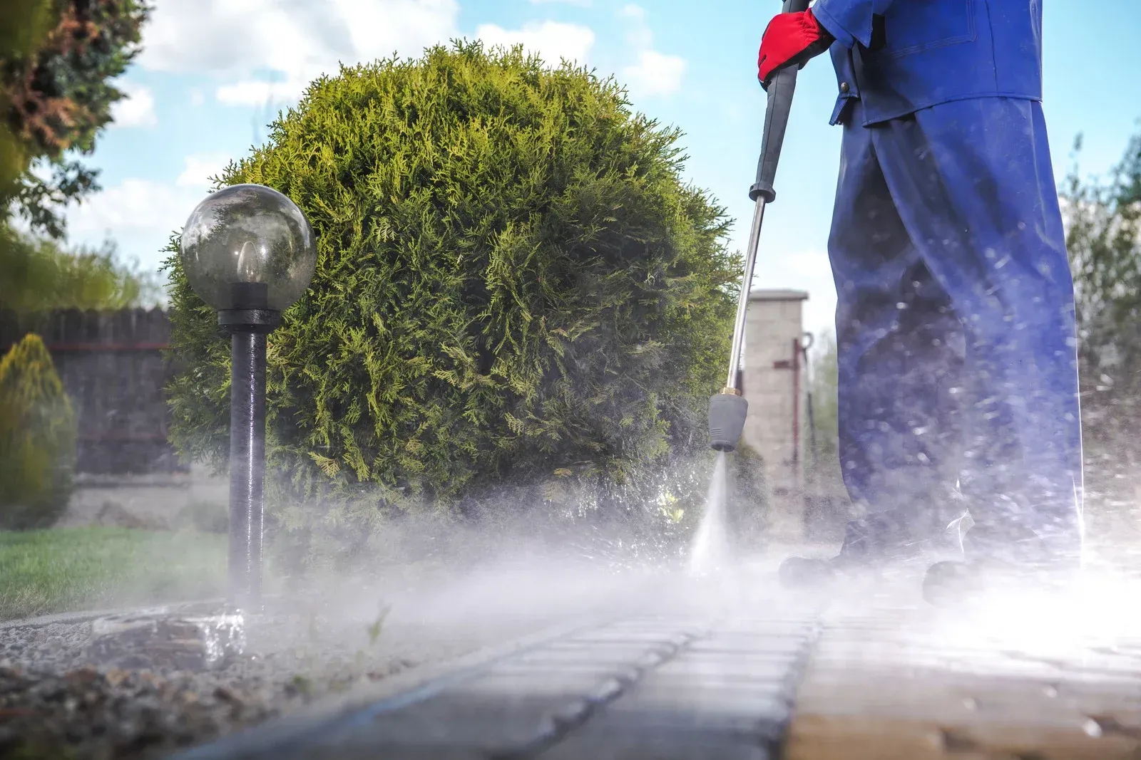 Person in blue overalls power washing a brick pathway, creating mist.