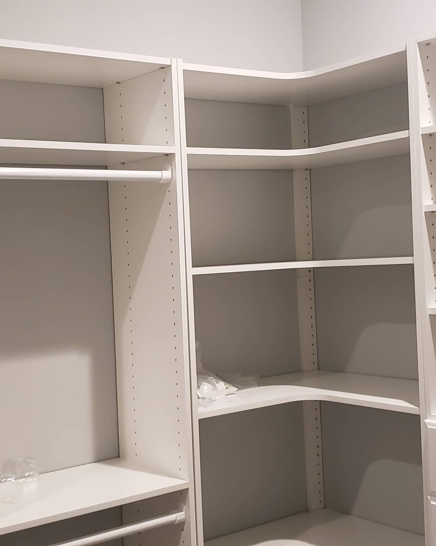 White built-in closet shelving system with hanging rod, shelves, and corner section. Empty, against a gray wall.