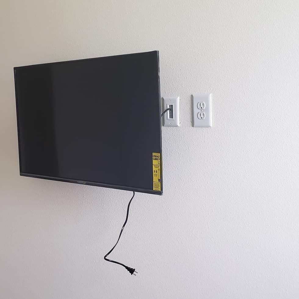 A TV mounted on a white wall. Next to it are electrical outlets. Black power cord hangs down.