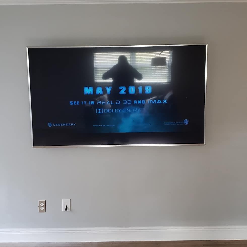Large TV screen on a gray wall, showing a movie trailer. Electrical outlets below the screen.