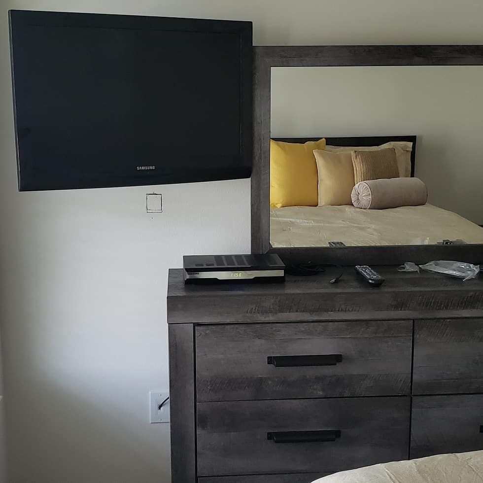 TV mounted on a white wall above a gray dresser with a mirror, reflecting a bed with pillows.