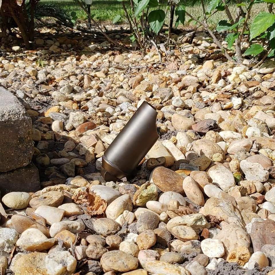 Brown metal outdoor landscape light on a bed of tan and brown rocks next to a stone border.