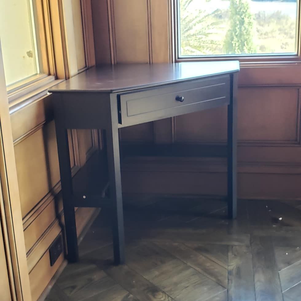 Dark wooden desk in a corner, with a single drawer and a window in the background.