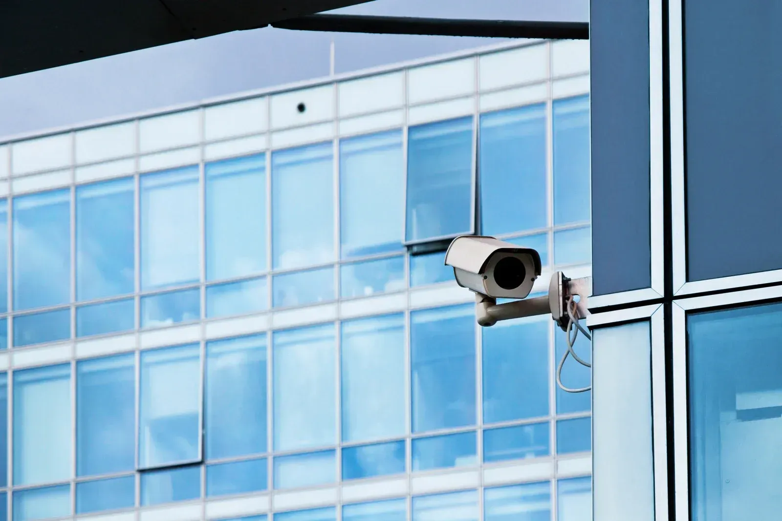 Security camera mounted on a modern building, overlooking a glass facade with blue windows.