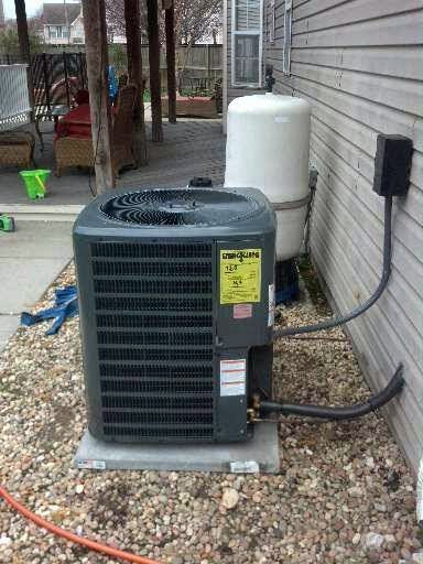 An outdoor HVAC unit next to a white water tank against a light-colored house wall, gravel ground.