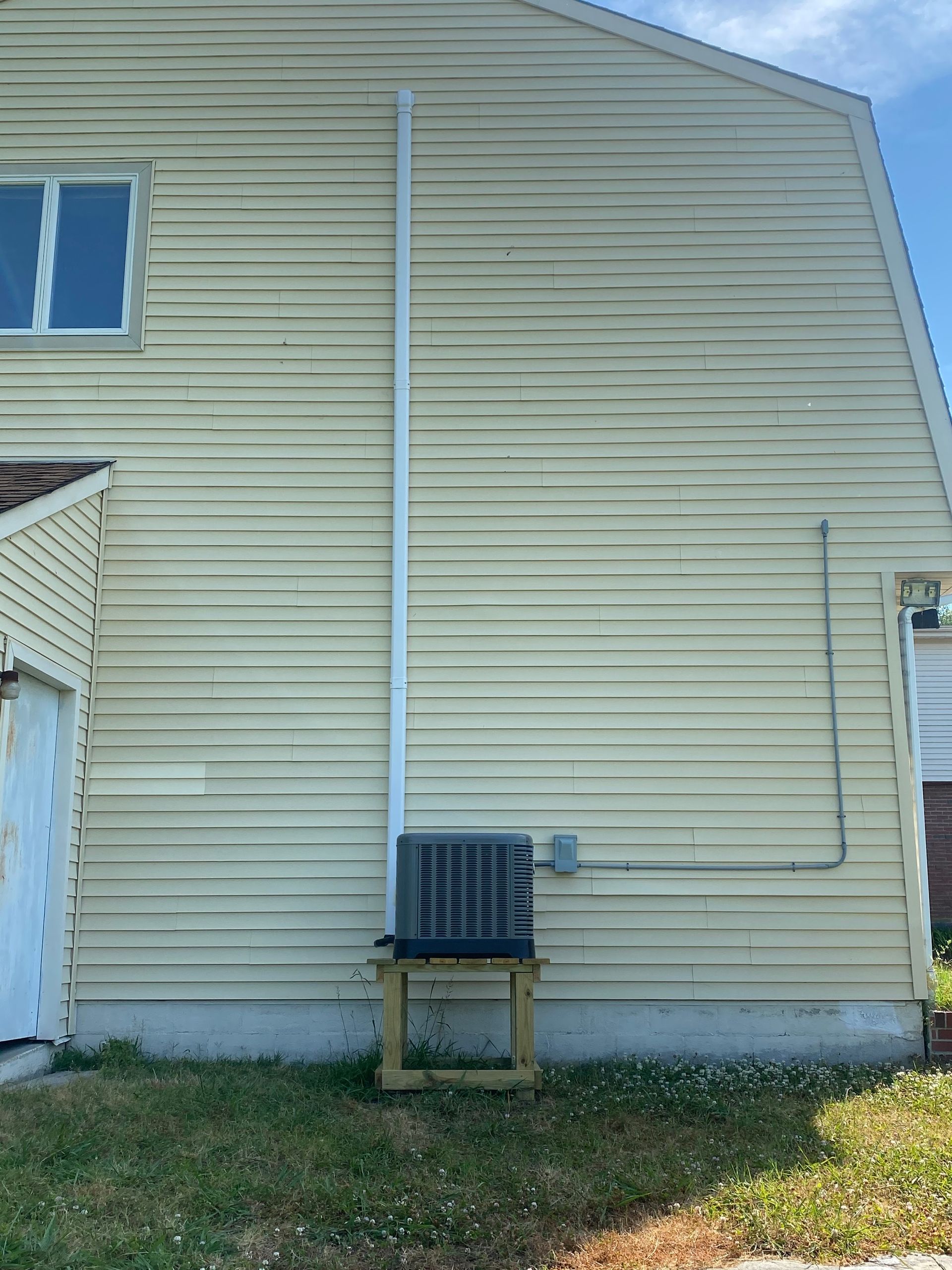 Exterior of a house with an air conditioning unit on a wooden stand. White siding and blue sky.