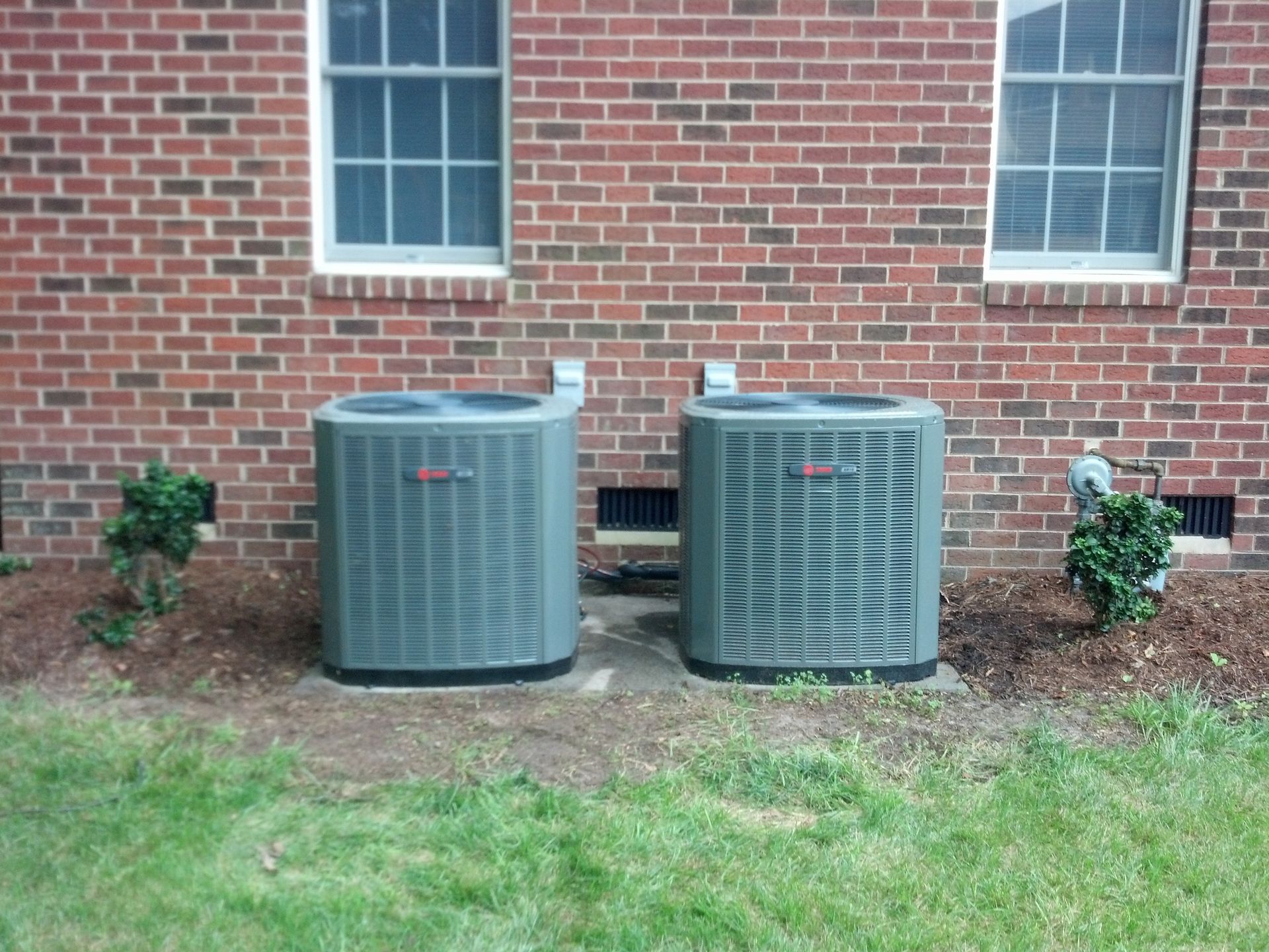 Two gray air conditioning units next to a brick building, with windows and small bushes.