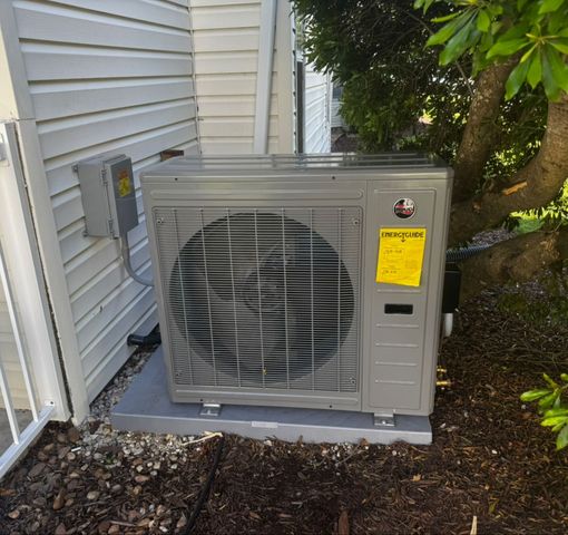 Outdoor air conditioning unit next to a white building and tree, mounted on a concrete slab.