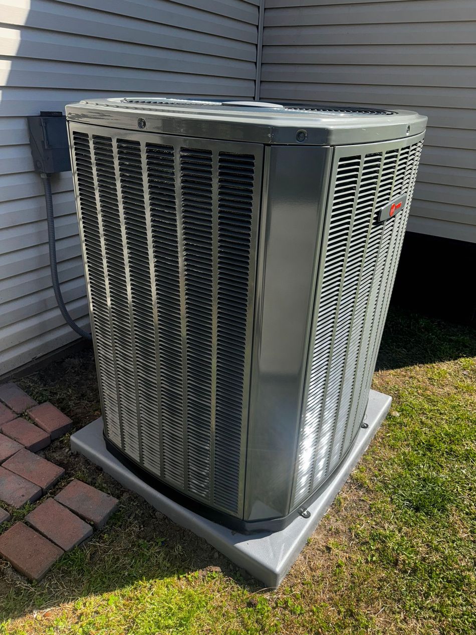 Gray air conditioning unit on a concrete pad, next to a house with brick and grass.