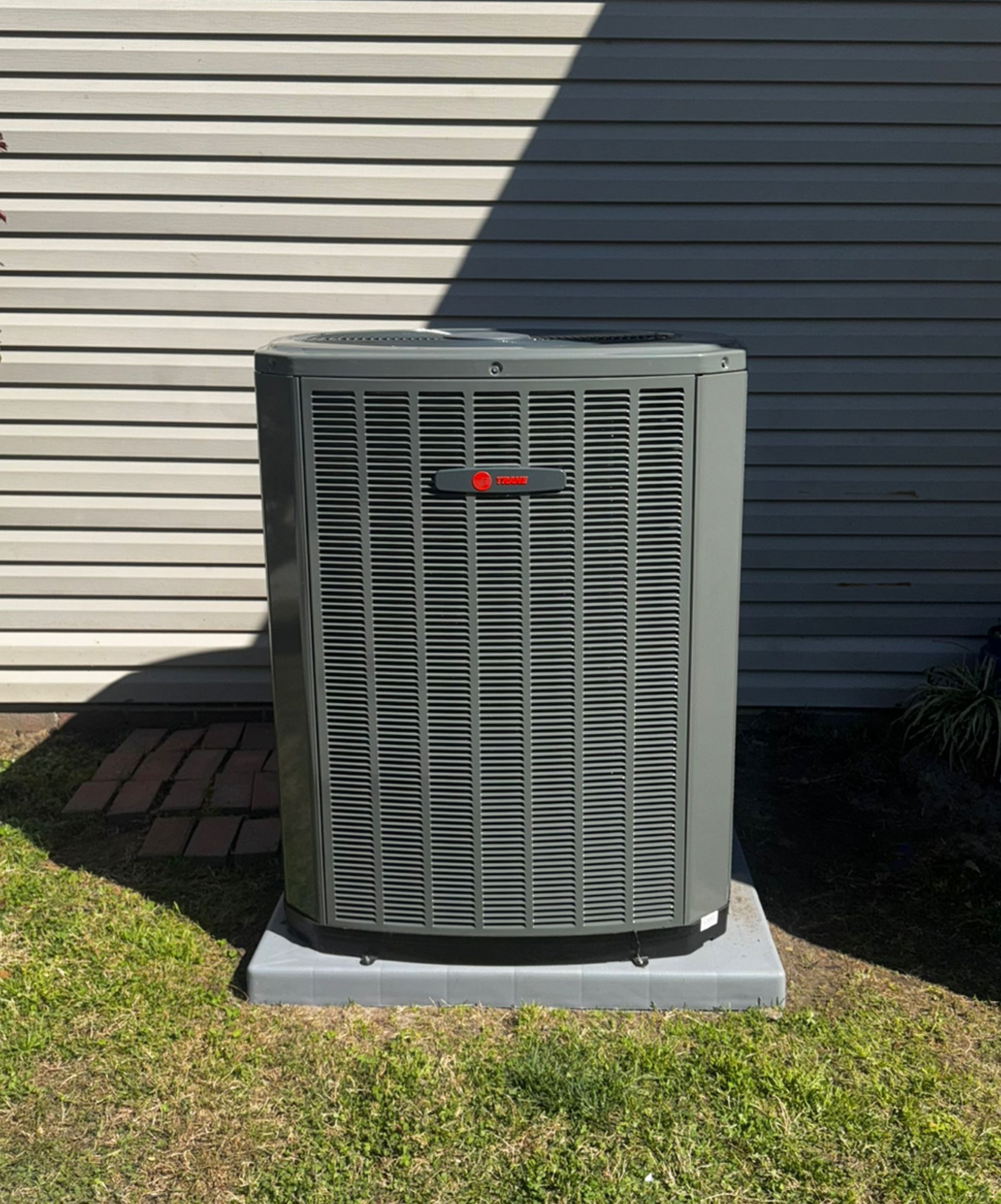 Gray Trane air conditioner unit on a concrete pad, in front of a light gray house wall.