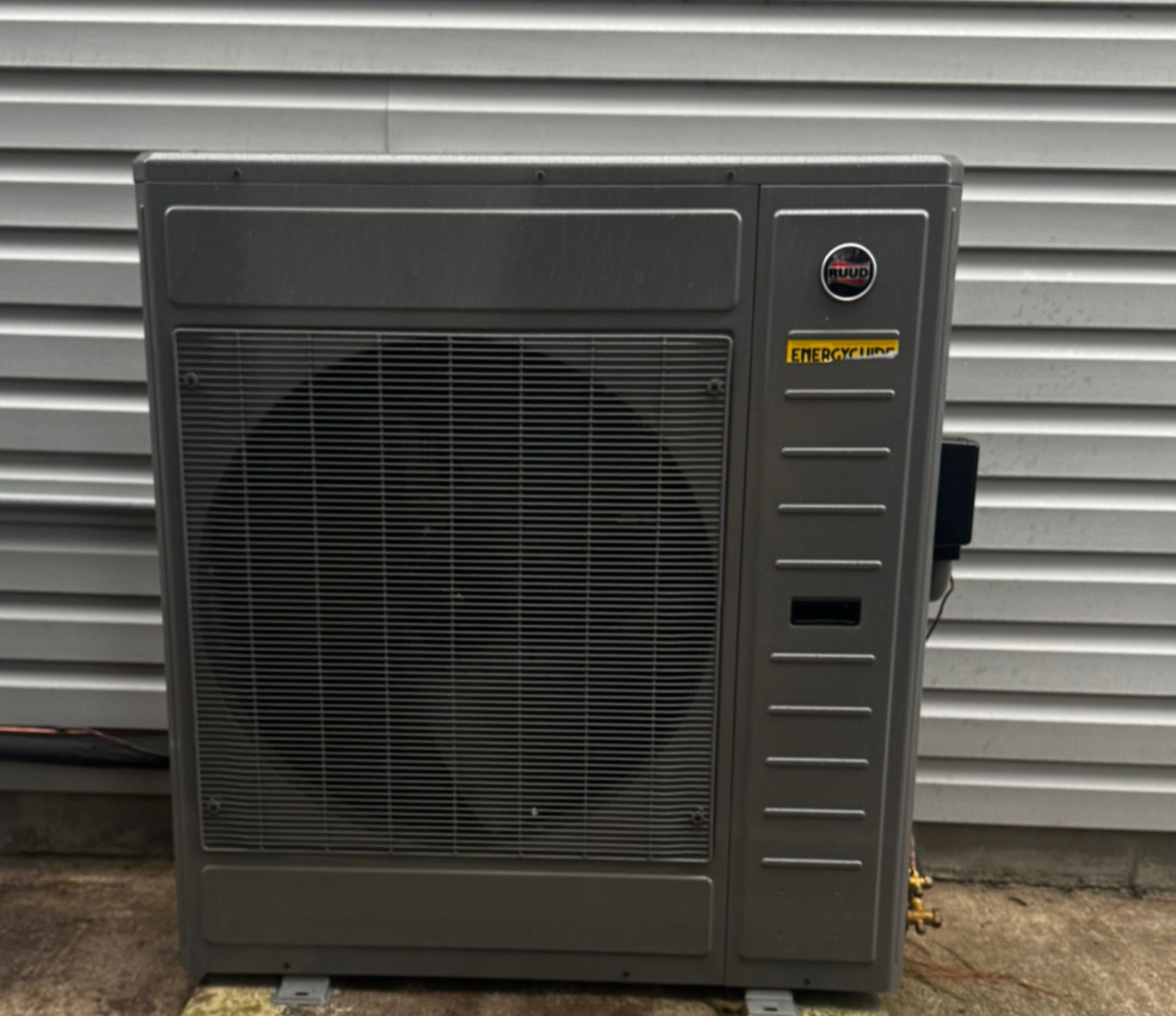 Gray HVAC unit with a large fan, mounted on a concrete pad, against a gray siding wall.