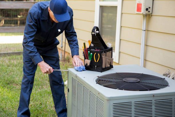 HVAC technician in blue overalls cleaning an outdoor AC unit with tools.