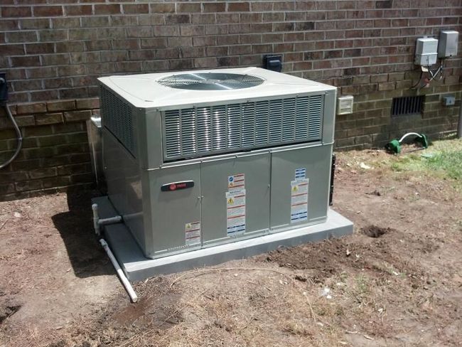 HVAC unit, silver and gray, on a concrete pad near a brick wall, with exposed ground in the foreground.