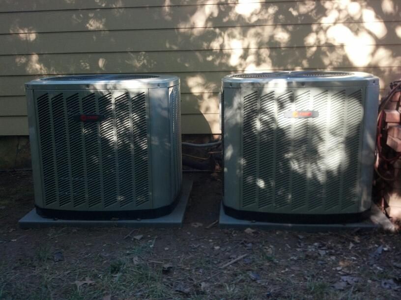 Outdoor air conditioning unit next to a white building and tree, mounted on a concrete slab.