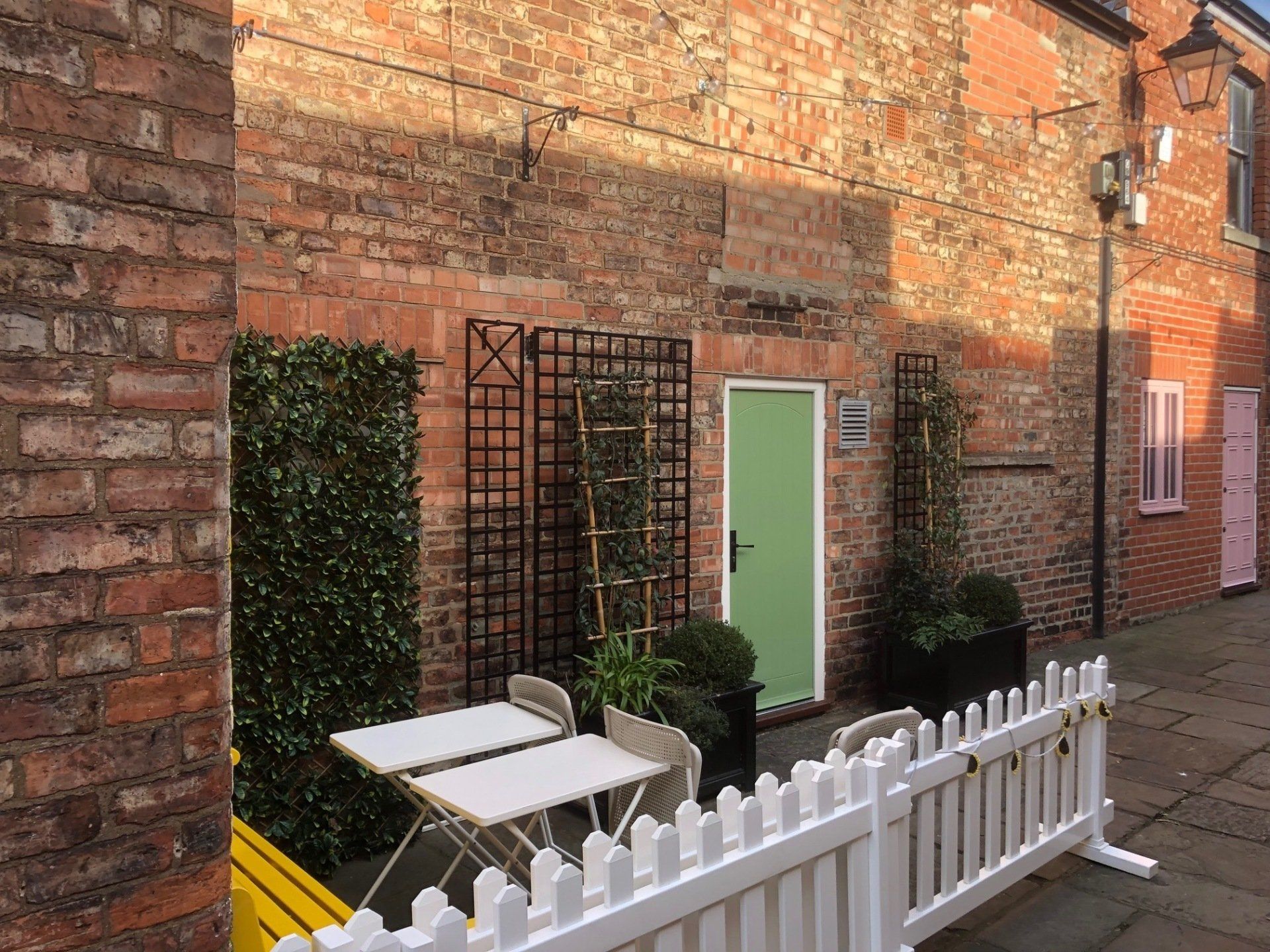 A white picket fence surrounds a brick building with tables and chairs in front of it.