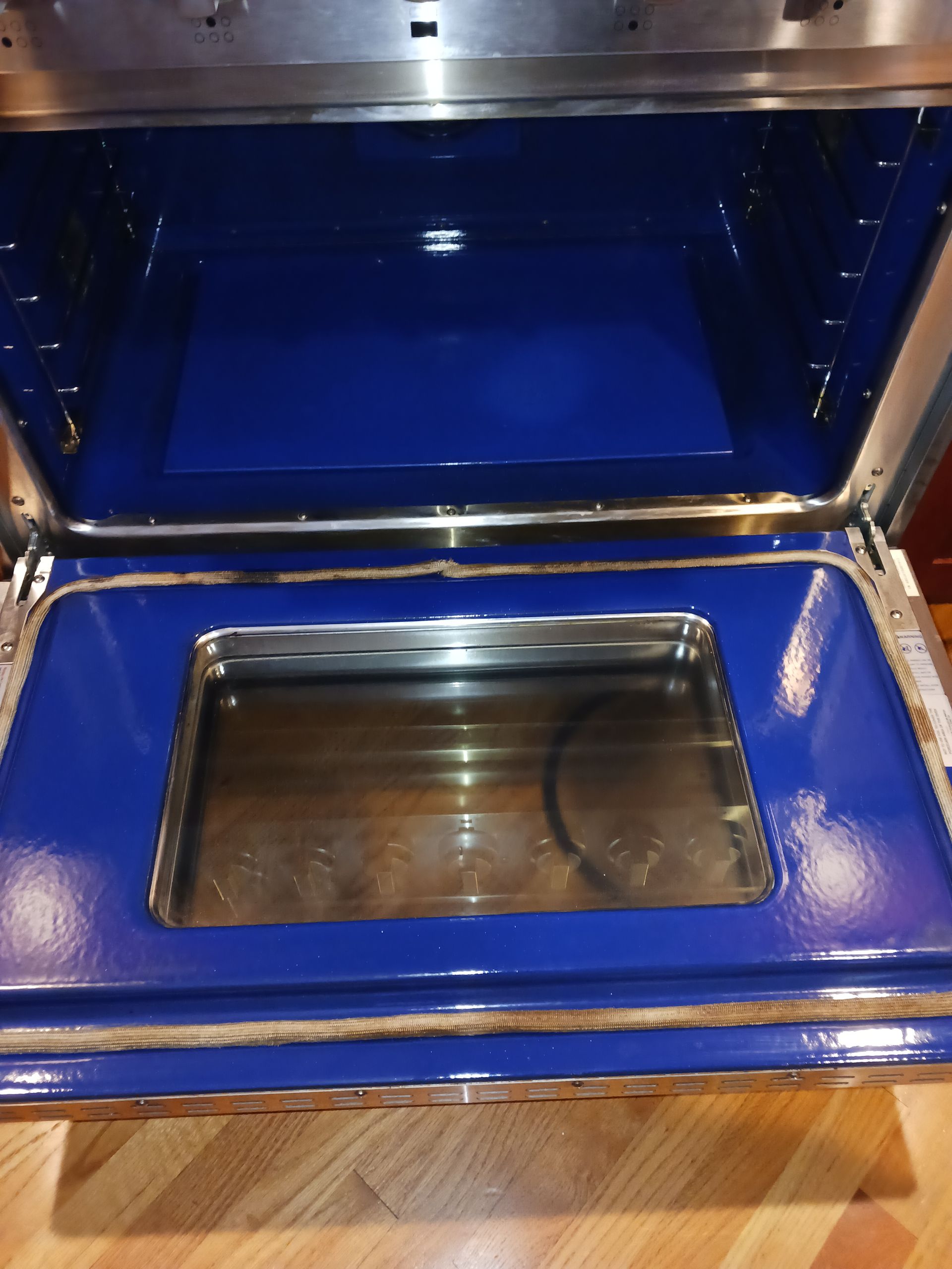 Open blue appliance with a stainless steel interior and glass viewing window on a wooden floor.