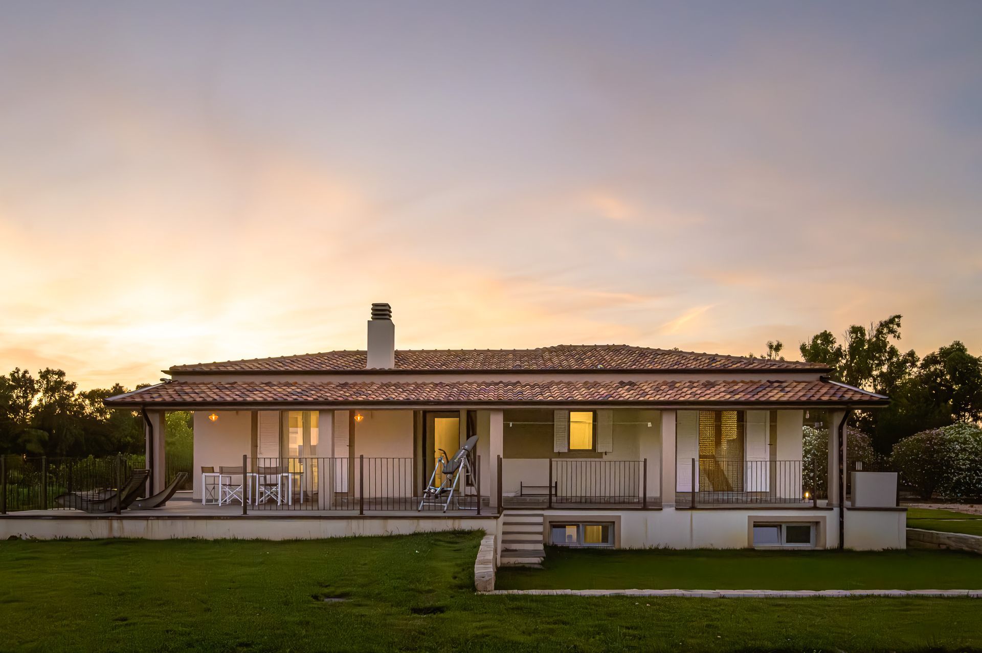 Cottage bianco con tetto, veranda e scale marroni. Prato verde e alberi con sfondo al tramonto.