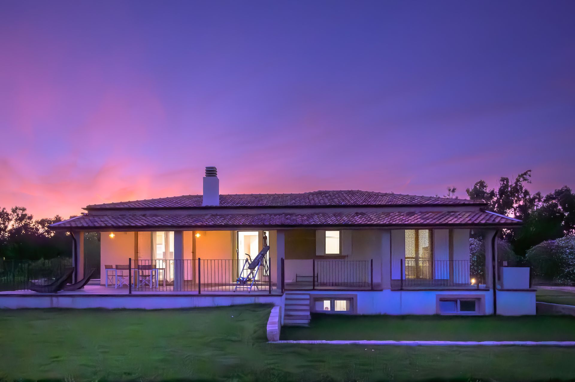 Cottage al tramonto con finestre illuminate e un cielo viola colorato. Prato verde di fronte.