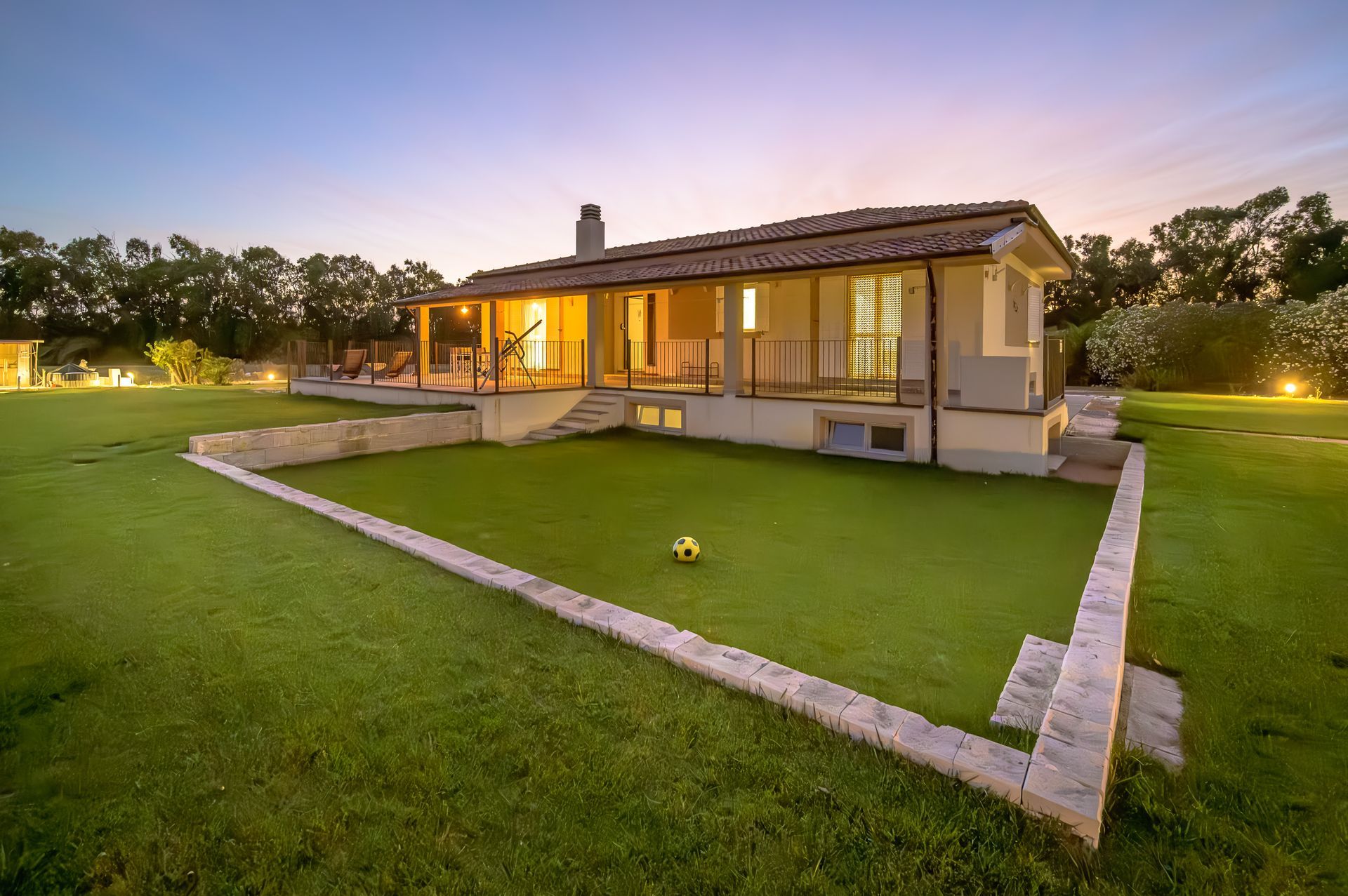 Una casa con un prato. Erba verde con un muro di pietra rialzato. Cielo al crepuscolo, con le luci della casa accese.