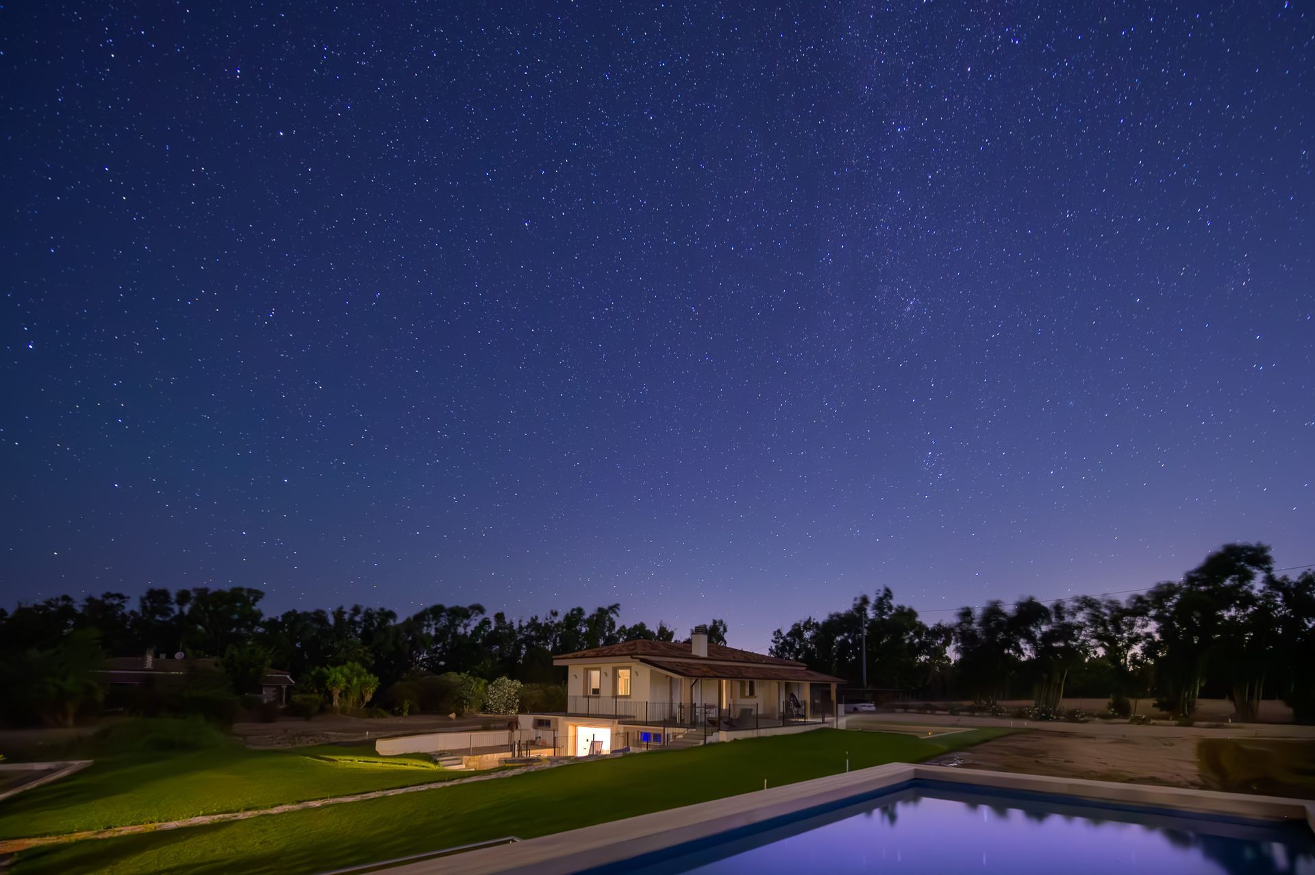Stelle sopra una casa di notte, cielo azzurro, luci accese, piscina, alberi in silhouette.