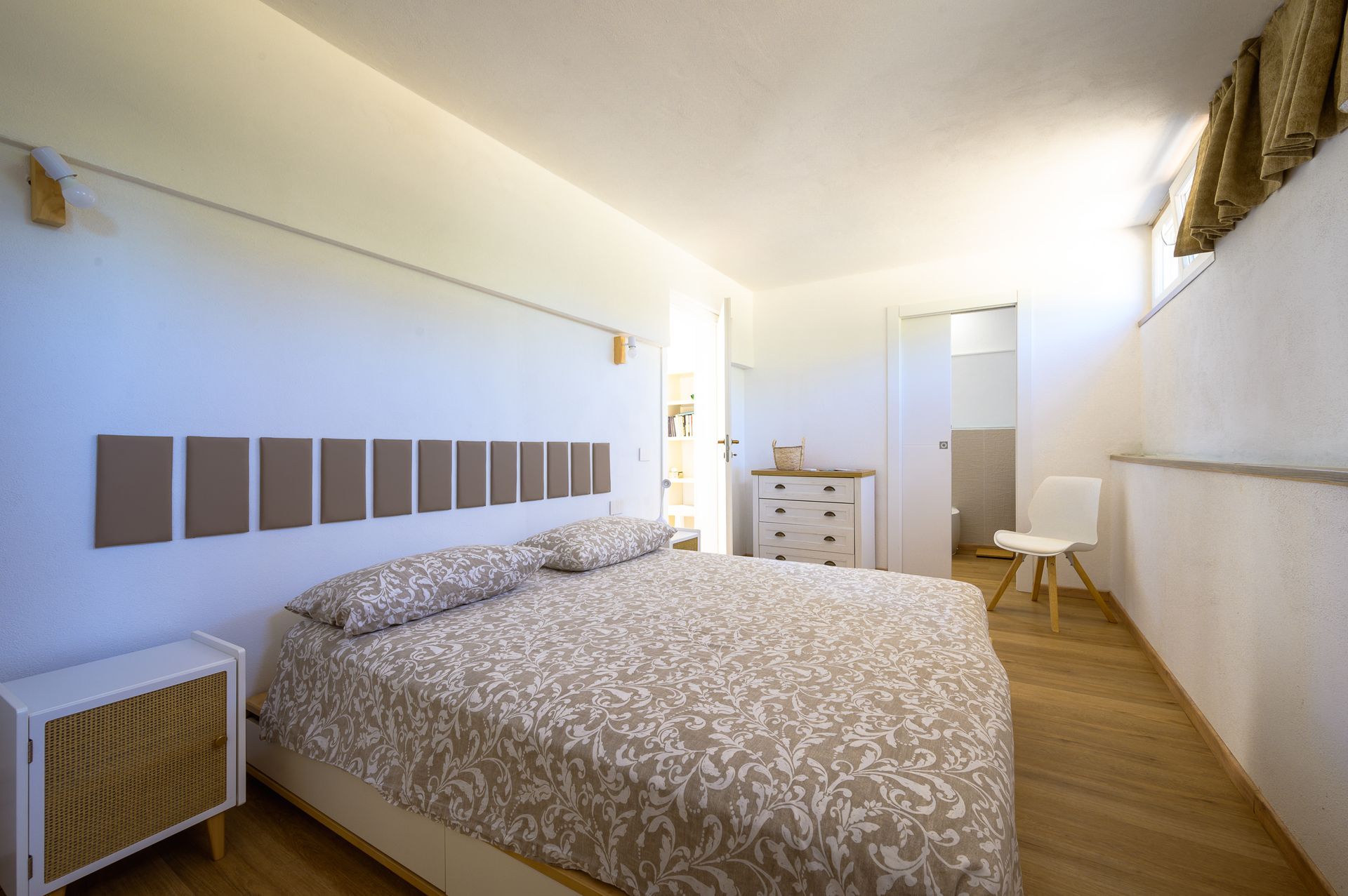 Camera da letto con letto, comodino, cassettiera e sedia. Pavimenti in legno chiaro e pareti bianche.