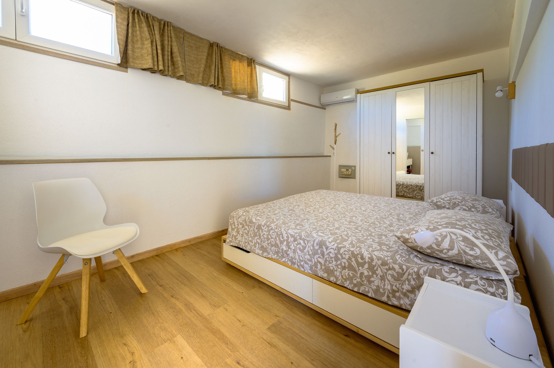 Camera da letto con letto, sedia bianca, armadio a specchio e piccole finestre. Pareti chiare e pavimento in legno.