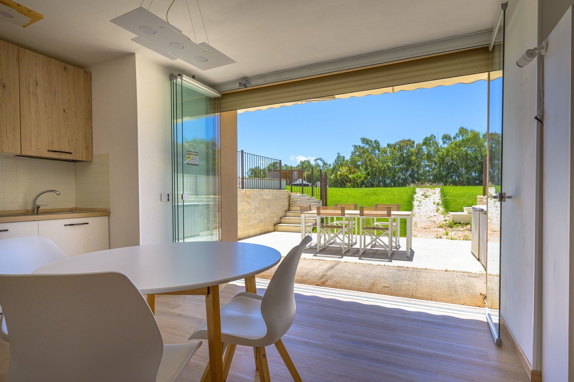 Vista interna con tavolo e sedie bianchi, porte a vetri aperte che conducono a una zona pranzo all'aperto.