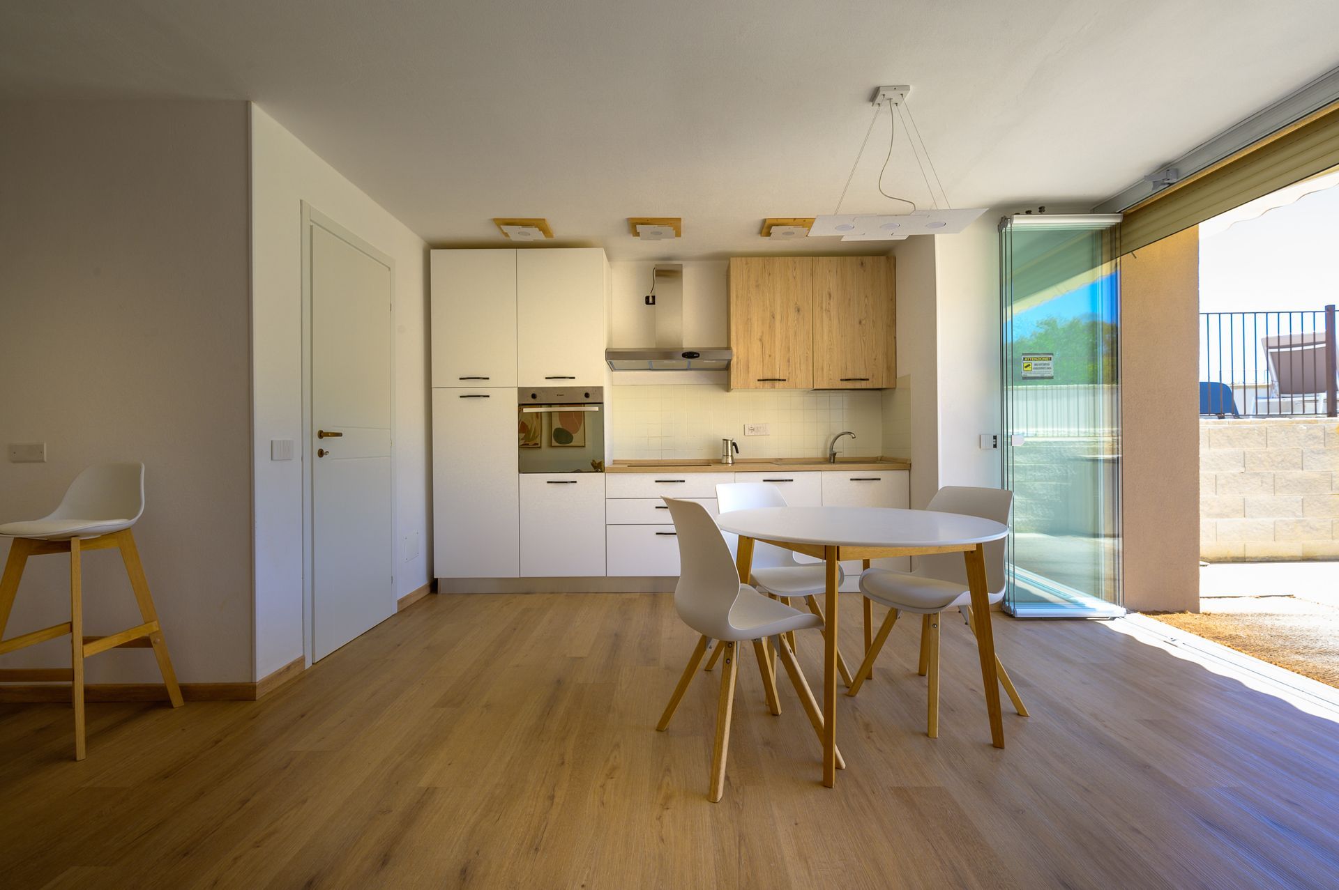 Cucina moderna con mobili bianchi e in legno, tavolo e sedie. Ampia porta a vetri che si apre sull'esterno.