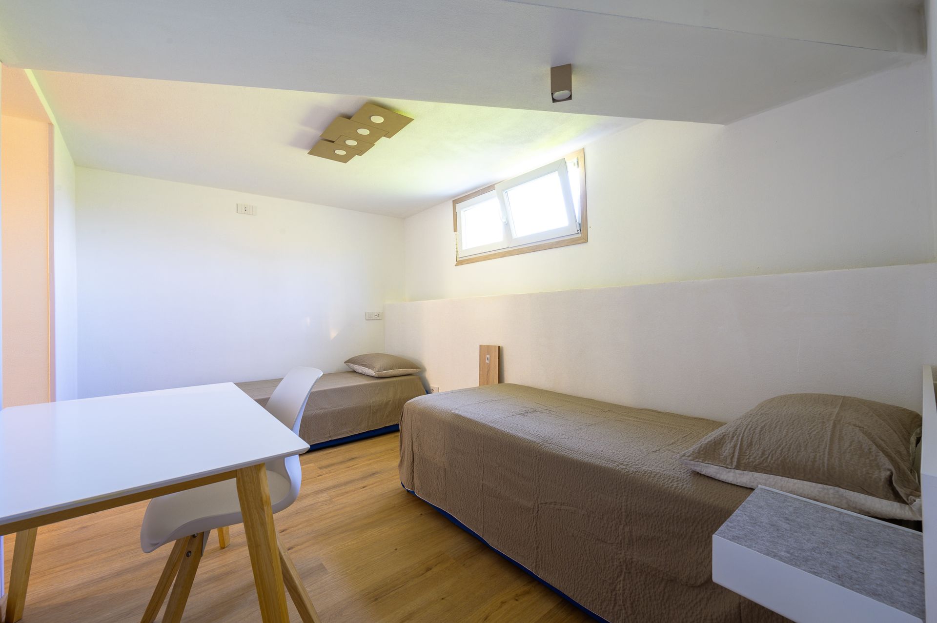 Camera da letto con due letti, un tavolo e una finestra, prevalentemente bianca e beige, con luce naturale.