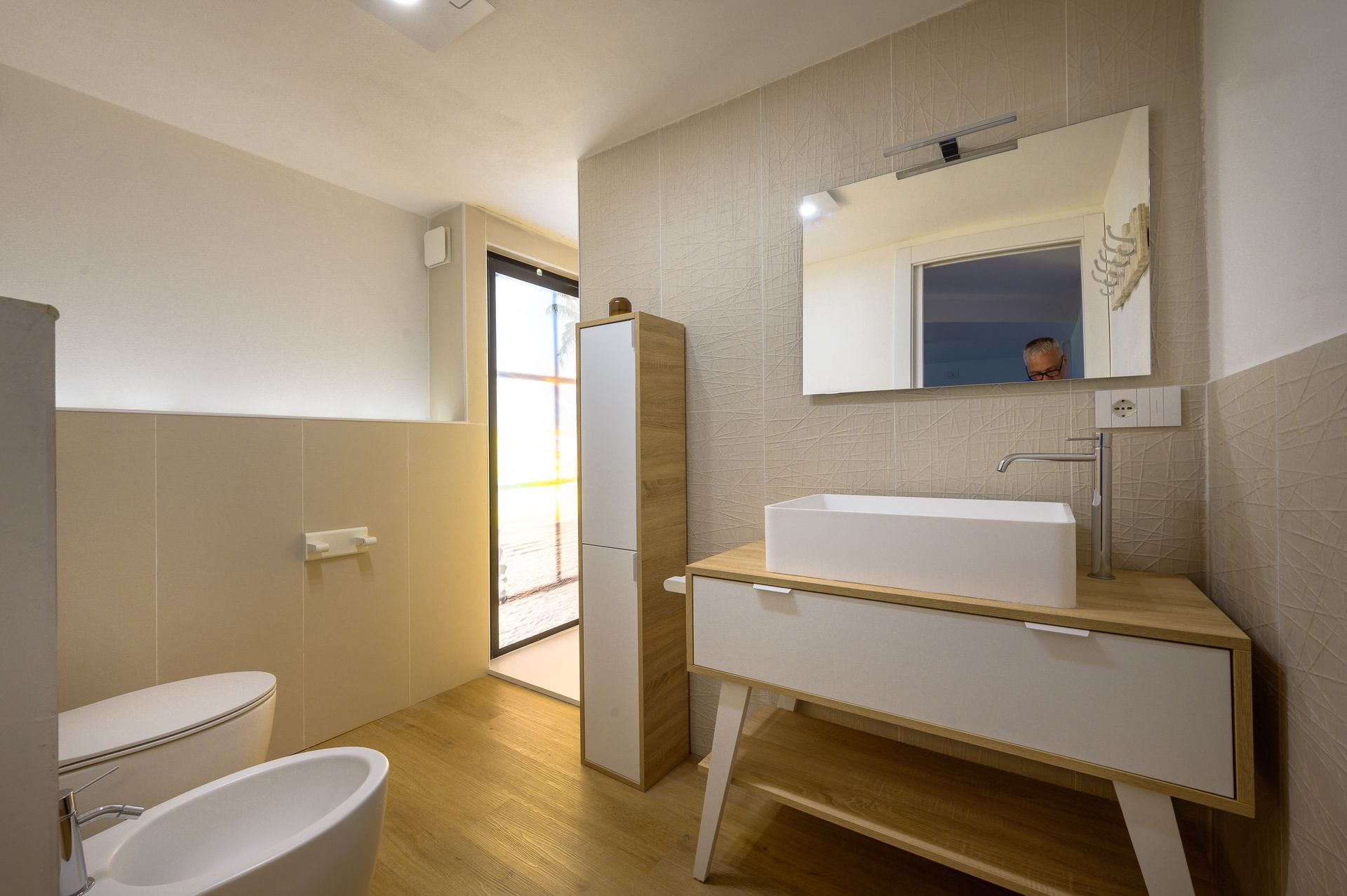 Bagno moderno con WC, lavandino e mobiletto bianchi; pavimenti in legno chiaro e pareti beige.