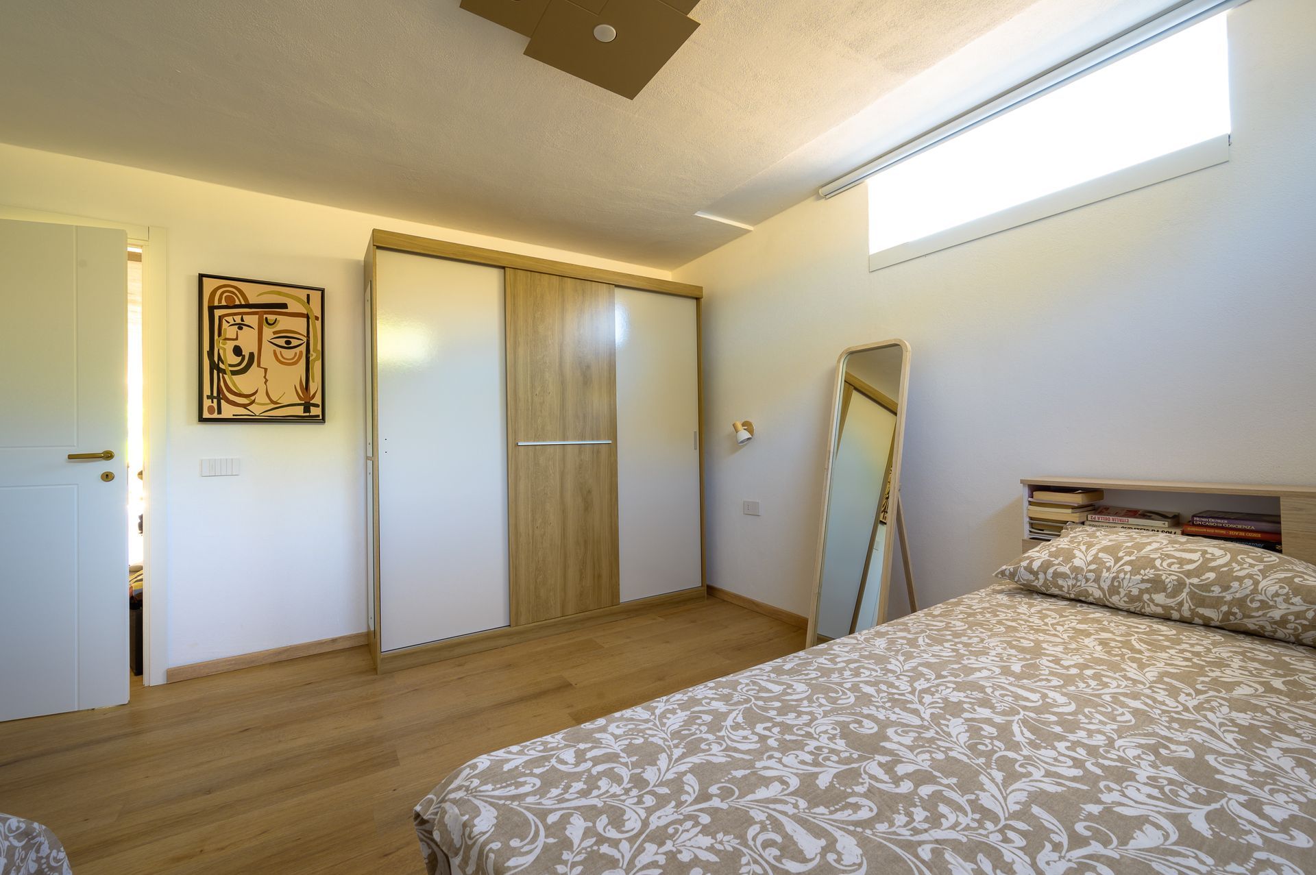 Camera da letto con letto, armadio, specchio a figura intera, porta aperta e opere d'arte su un pavimento in legno chiaro.