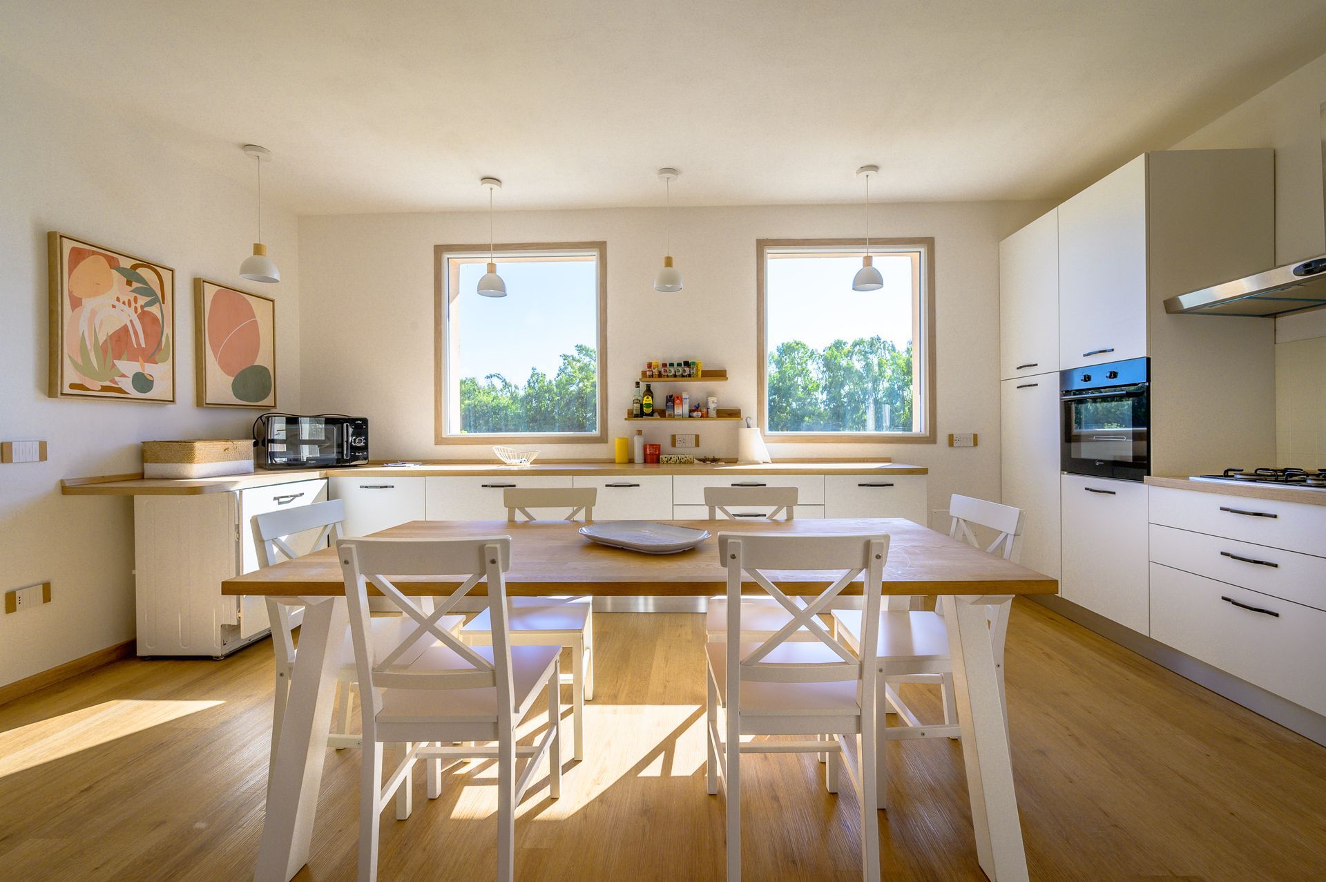 Cucina luminosa con tavolo da pranzo in legno, sedie bianche, mobili e finestre con vista sugli alberi verdi.