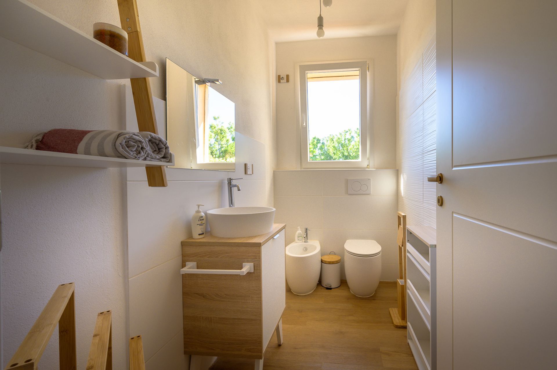 Un bagno bianco luminoso con lavandino, WC e finestra, con dettagli in legno e mensole.