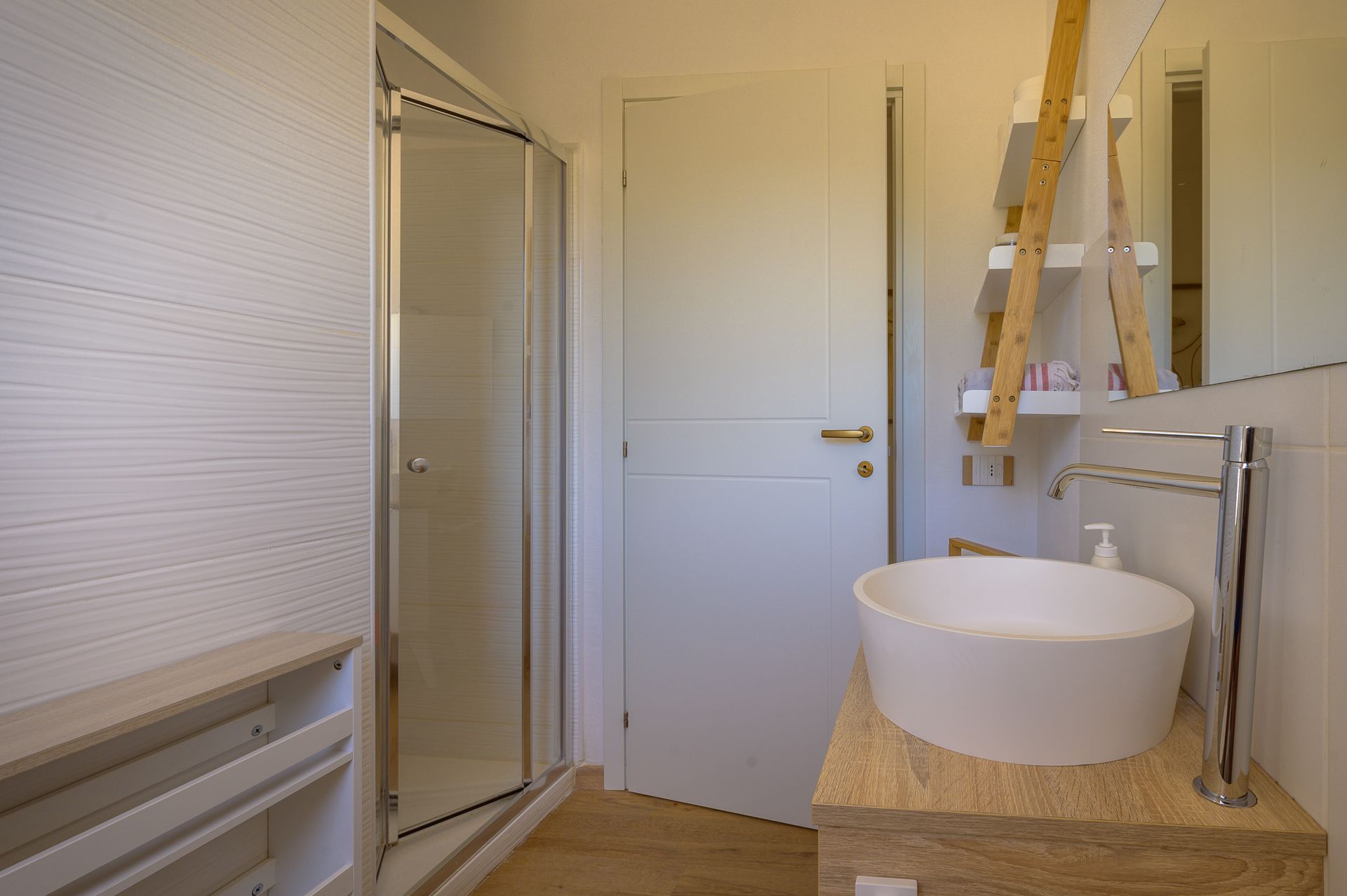 Bagno moderno con doccia, lavandino e porta; pareti chiare, ripiani in legno e ripiano in pietra.