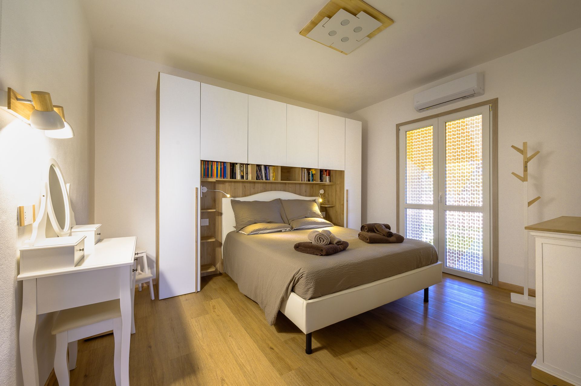 Camera da letto con letto, armadio bianco con libreria, toletta e finestra. Pavimento e pareti in legno.