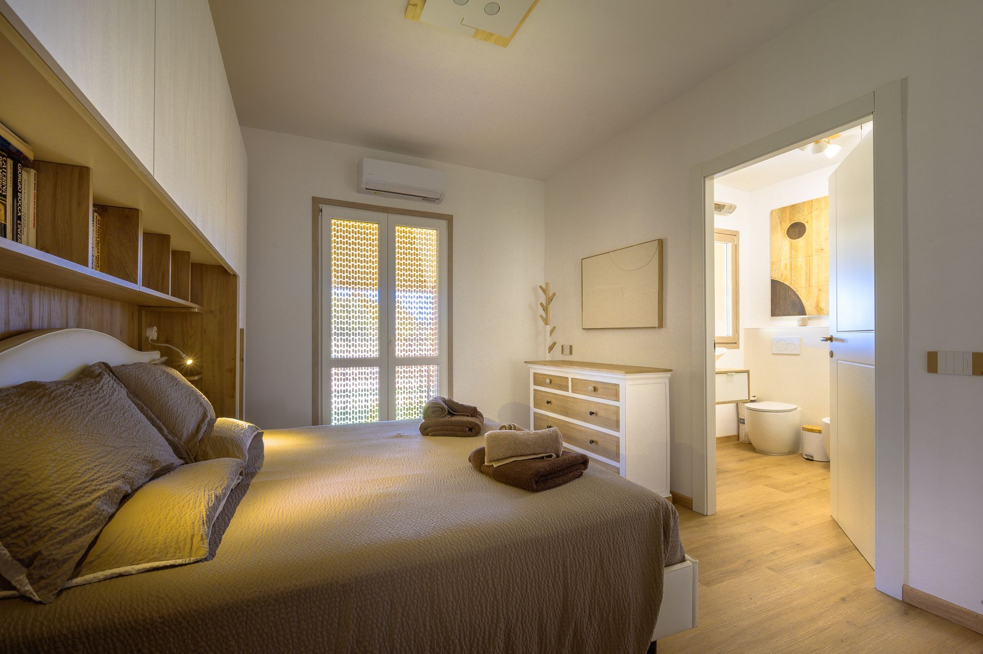 Camera da letto con letto, cassettiera, porta aperta sul bagno e finestra con luce solare.