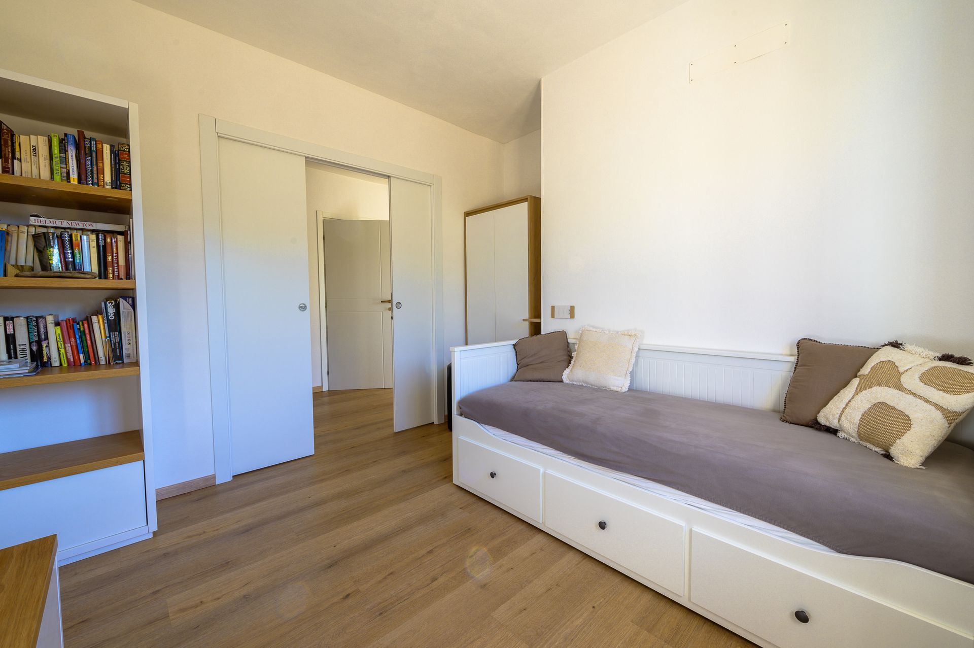 Camera da letto con divano letto bianco, libreria con libri e porta aperta su un'altra stanza.