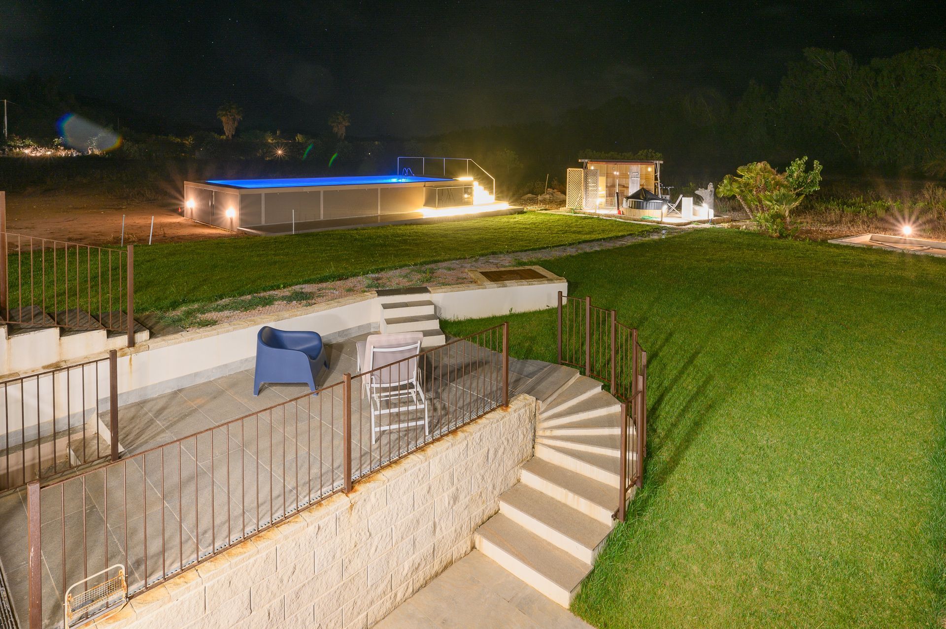 Vista notturna di un cortile con piscina, patio e prato erboso, illuminato da luci.