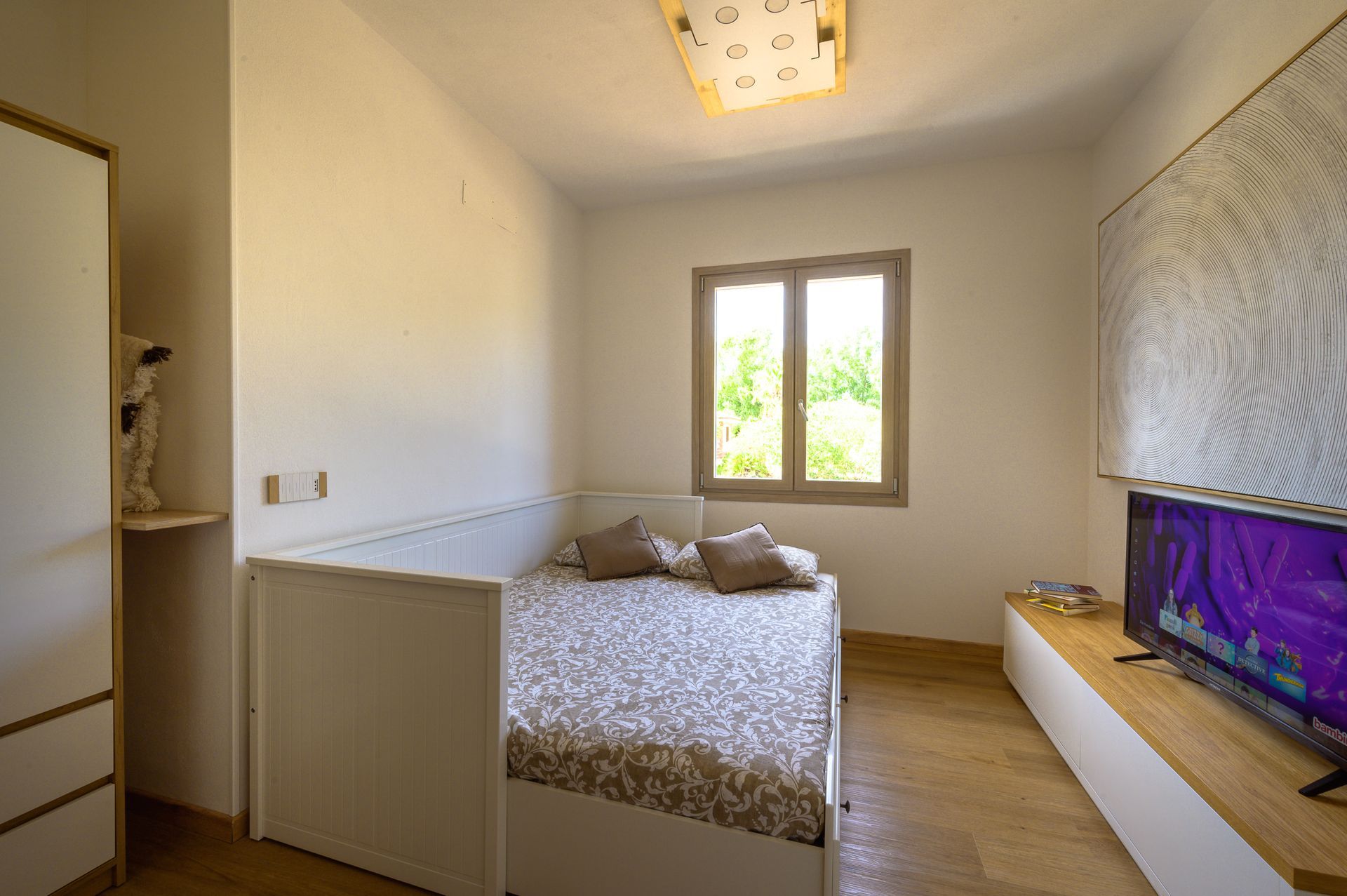 Una camera da letto con letto, finestra, TV e armadio. La luce naturale inonda la stanza.