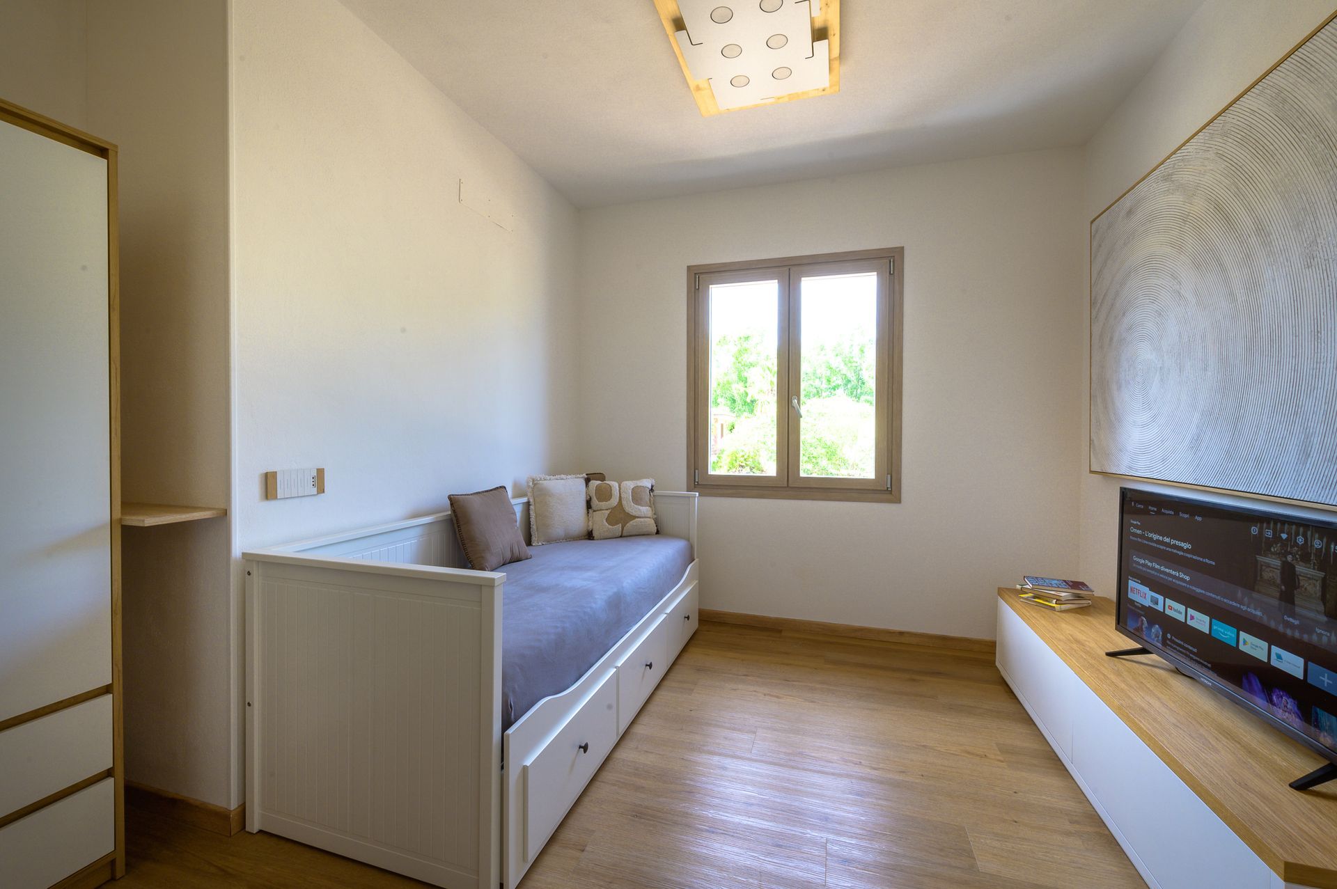 Camera da letto con divano letto, TV su una mensola sospesa e finestra che lascia entrare la luce naturale.