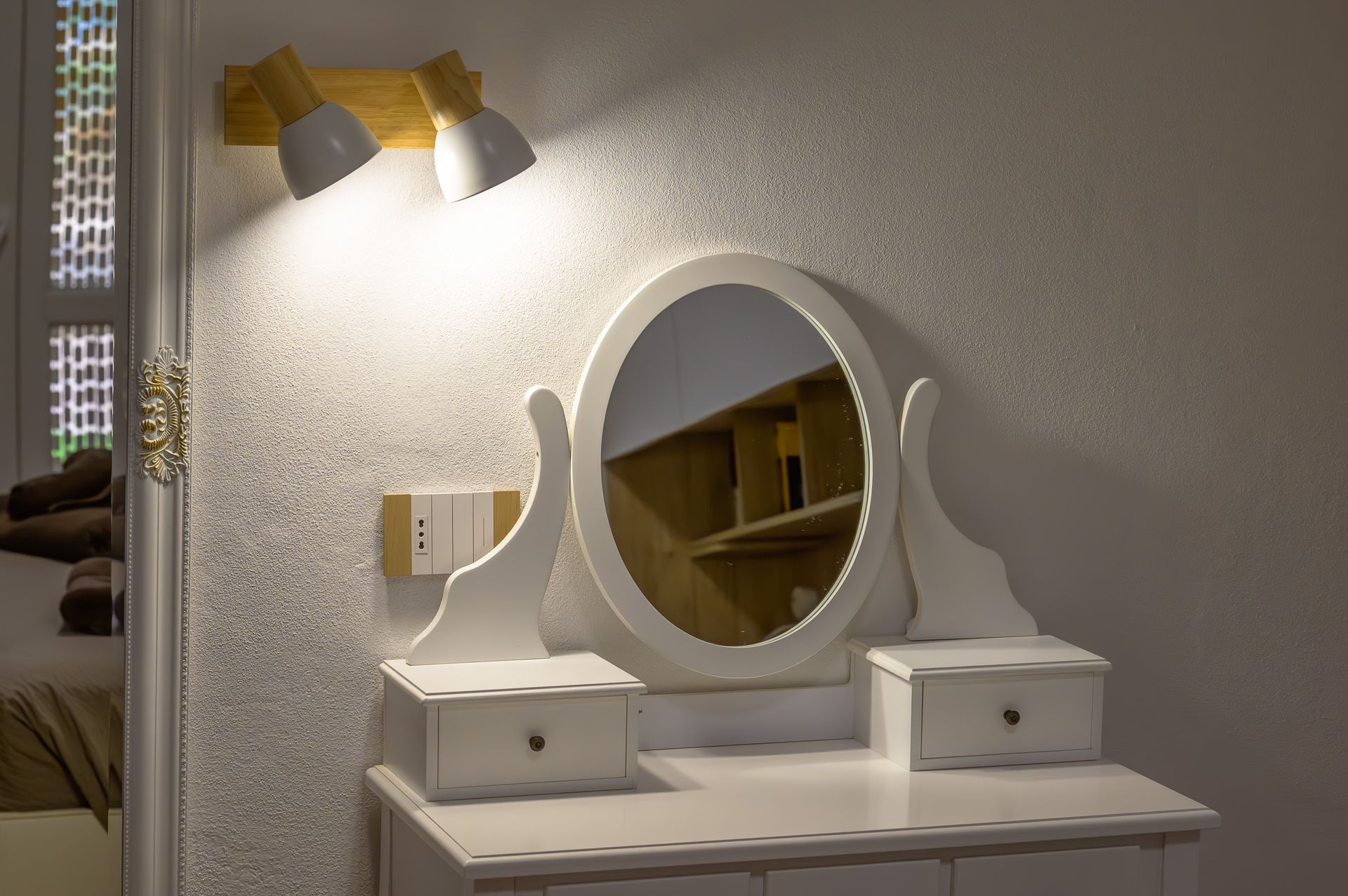 Tavolo da toeletta bianco con specchio ovale e due cassetti, illuminato da una lampada a parete.