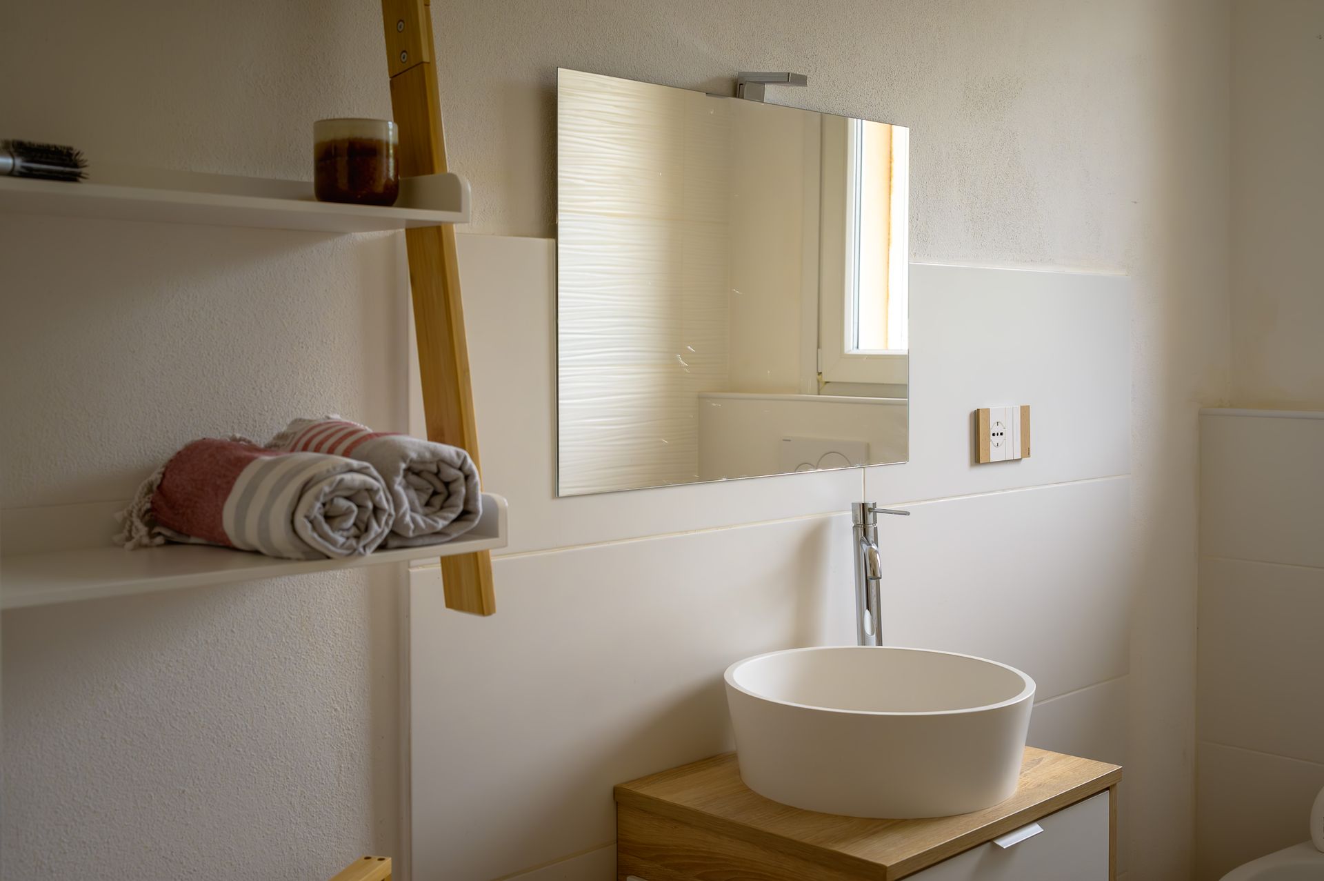 Bagno con lavandino bianco, specchio, asciugamani e mensole. Dettagli in legno.