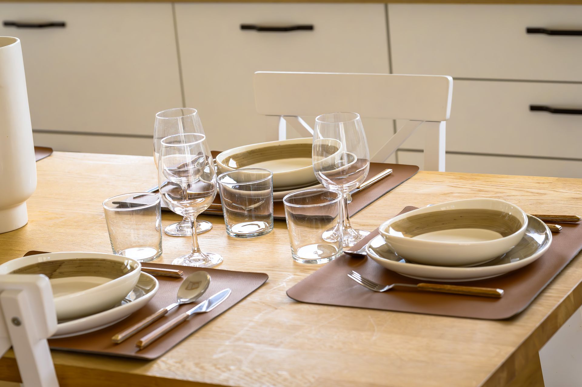 Tavolo da pranzo apparecchiato con piatti, bicchieri, posate su tovagliette marroni.