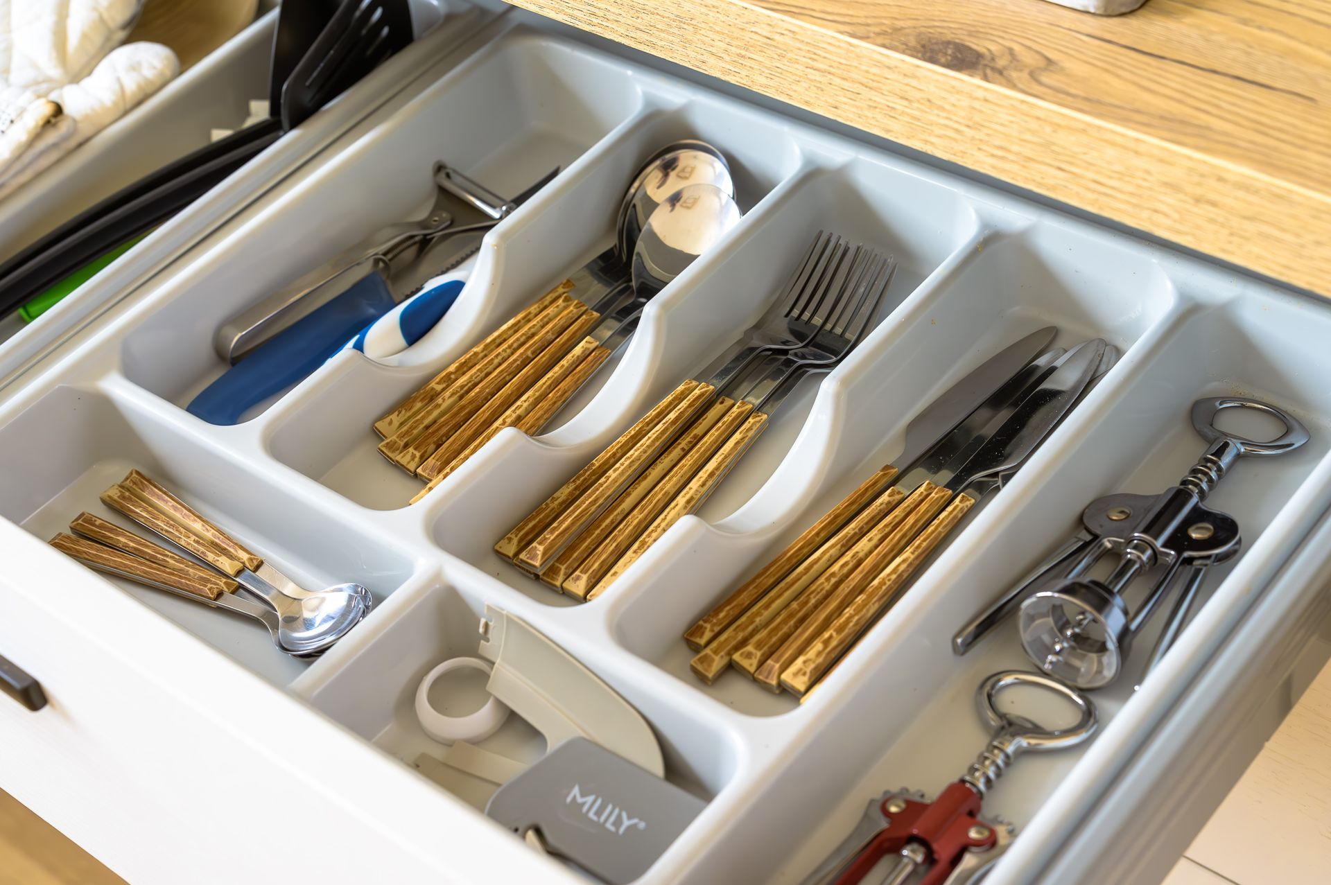 Cassetto delle posate con posate in argento, utensili con manico dorato, apribottiglie e divisori bianchi per cassetti.