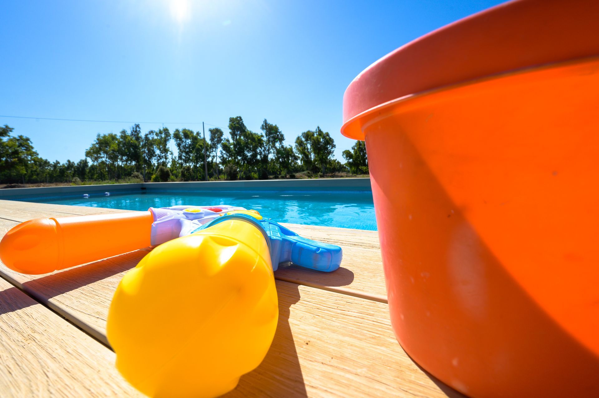Scena a bordo piscina con giocattoli: secchio arancione, pistole ad acqua e acqua blu contro un cielo luminoso.