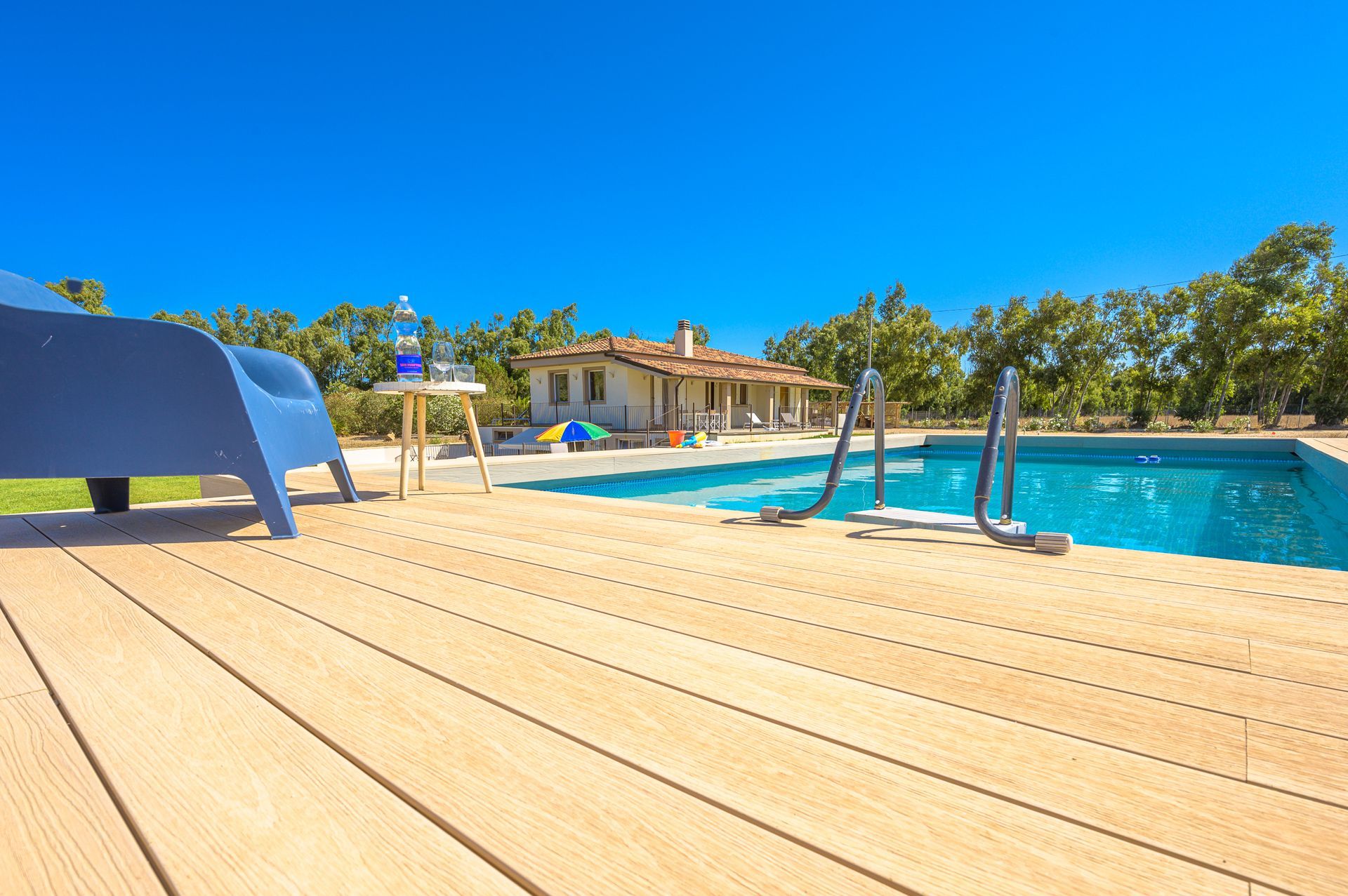 Scena a bordo piscina: sedia blu su terrazza in legno, piscina, tavolino, casa, cielo sereno.