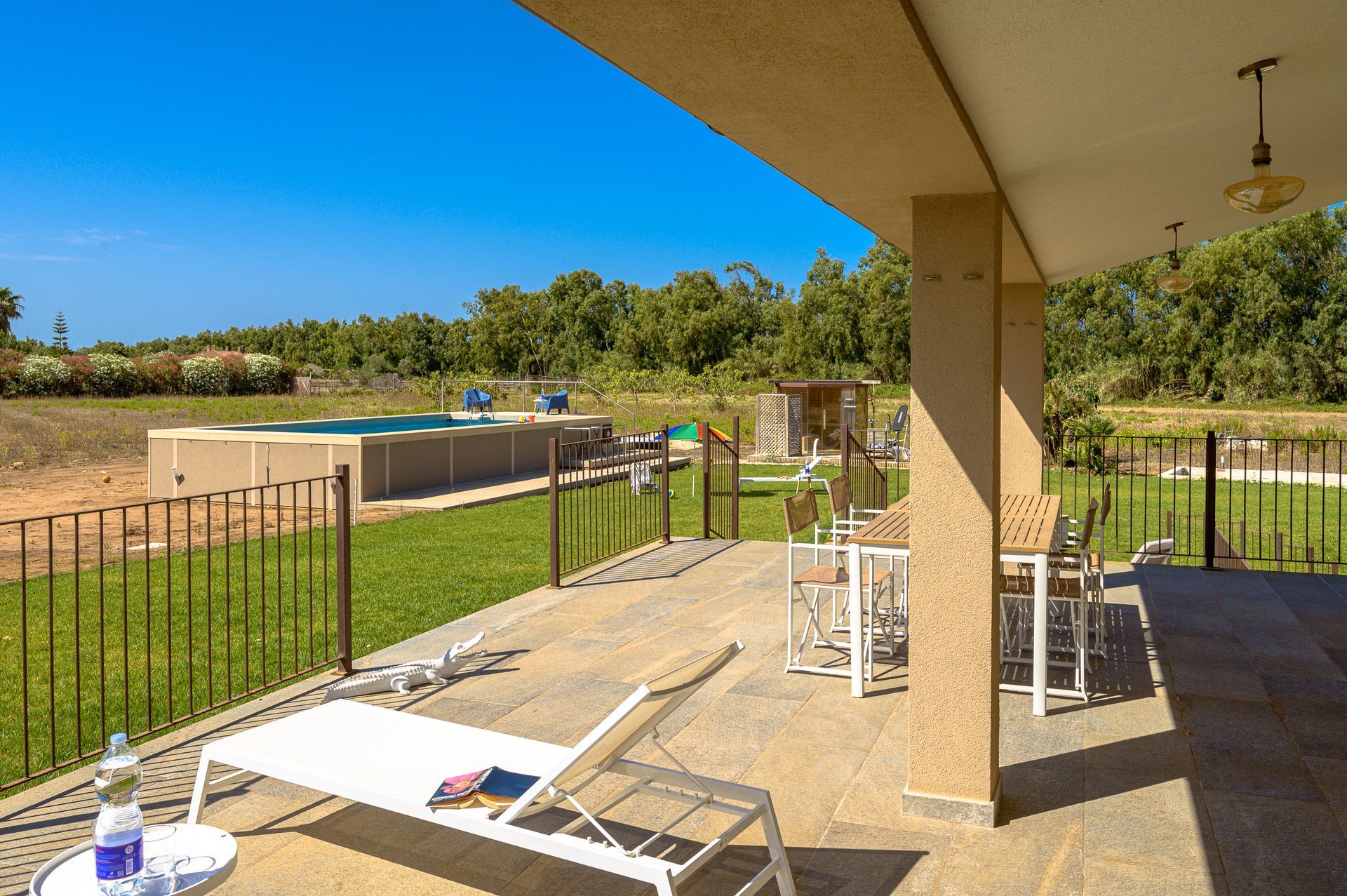 Patio con vista sulla piscina e sul prato sotto un cielo azzurro. Sono visibili una chaise longue e delle sedie.